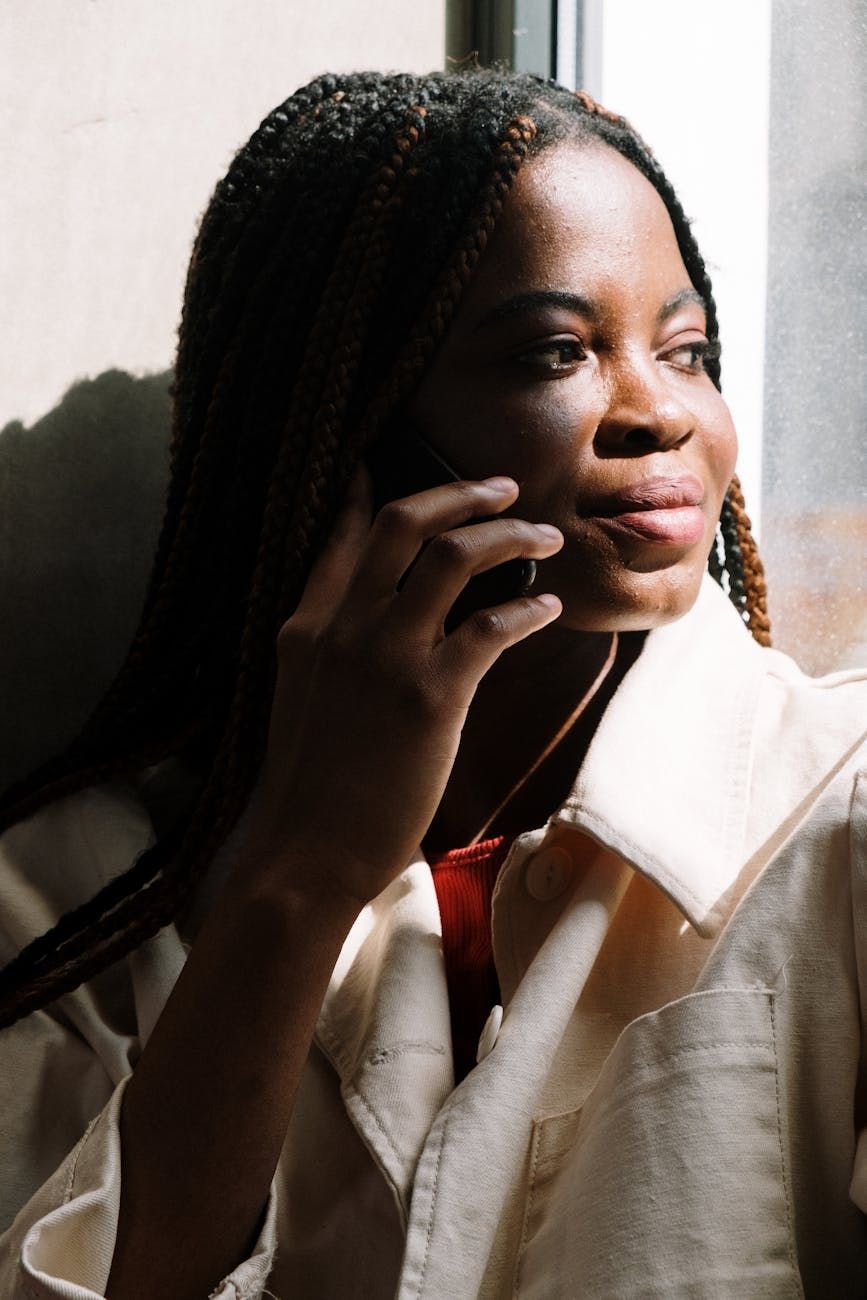 A woman with braided hair talking on a cellphone by a sunny window, warm morning light.