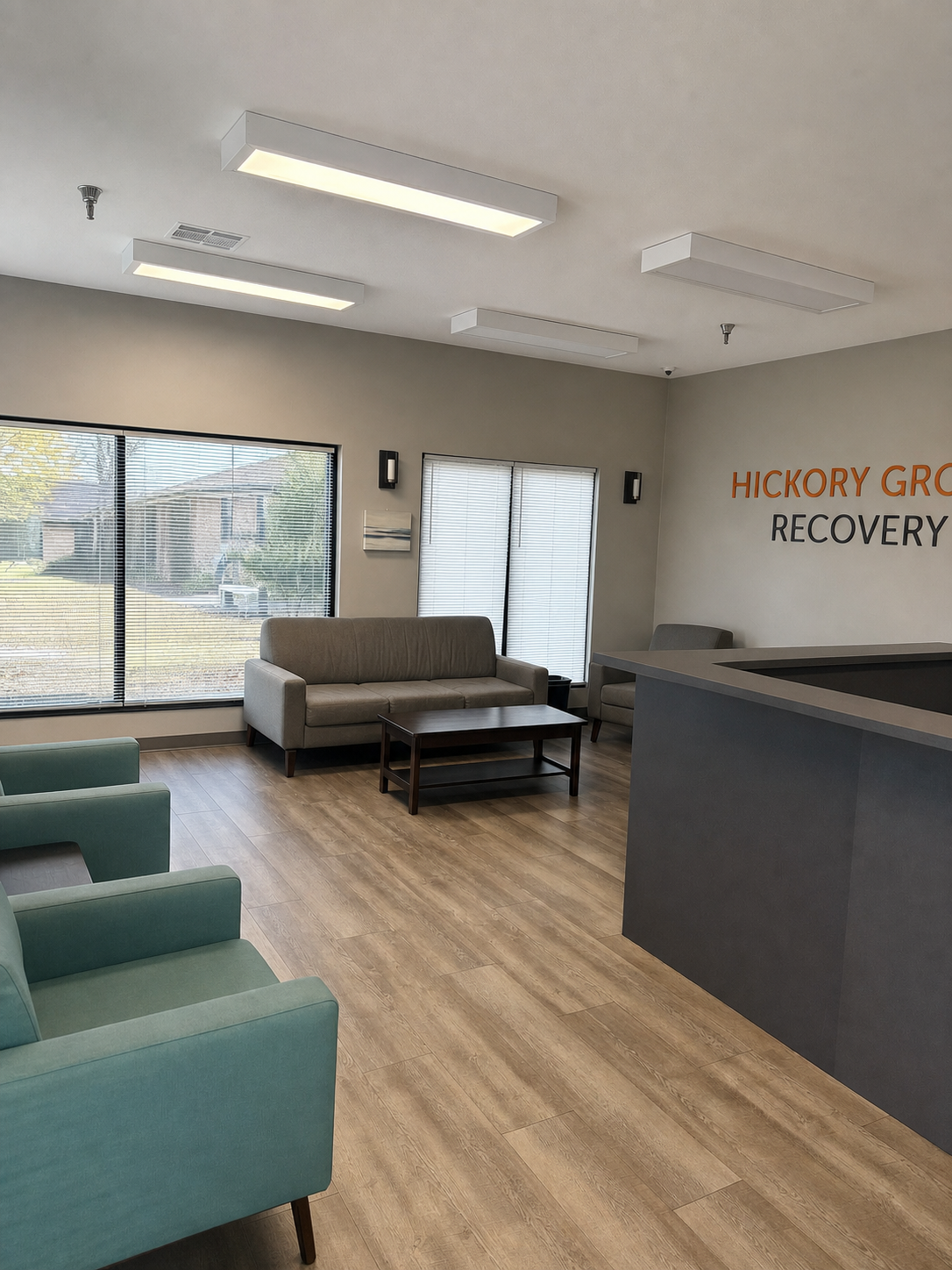 The lobby at Hickory Grove Recovery in Morrilton, Arkansas — sofa and sage armchairs by tall windows, with the Hickory Grove Recovery wordmark on the wall.