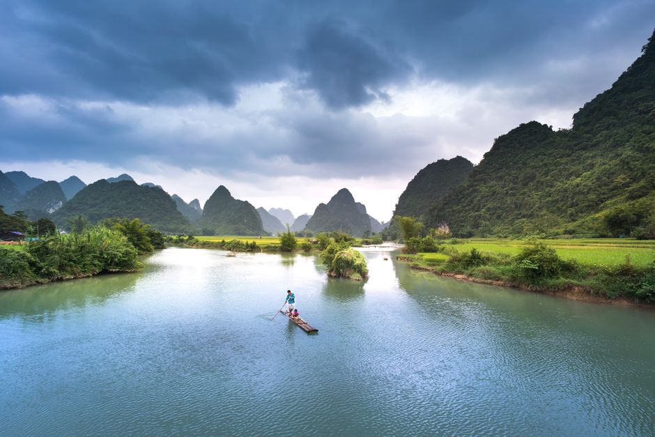 A serene view of a river with lush green mountains under a dramatic sky.