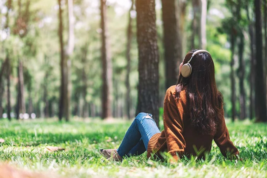 a woman sits in the grass with headphones on in a wooded area