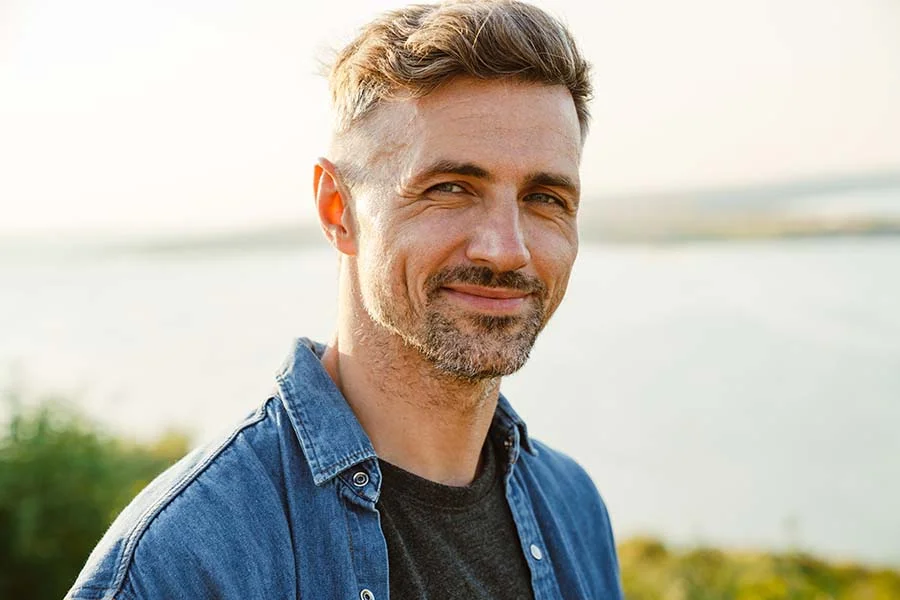a smiling man looks at the camera with a lake behind him
