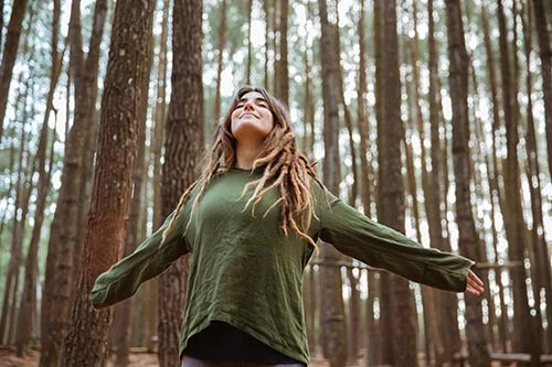 a woman stands with arms outstretched in a wooded area